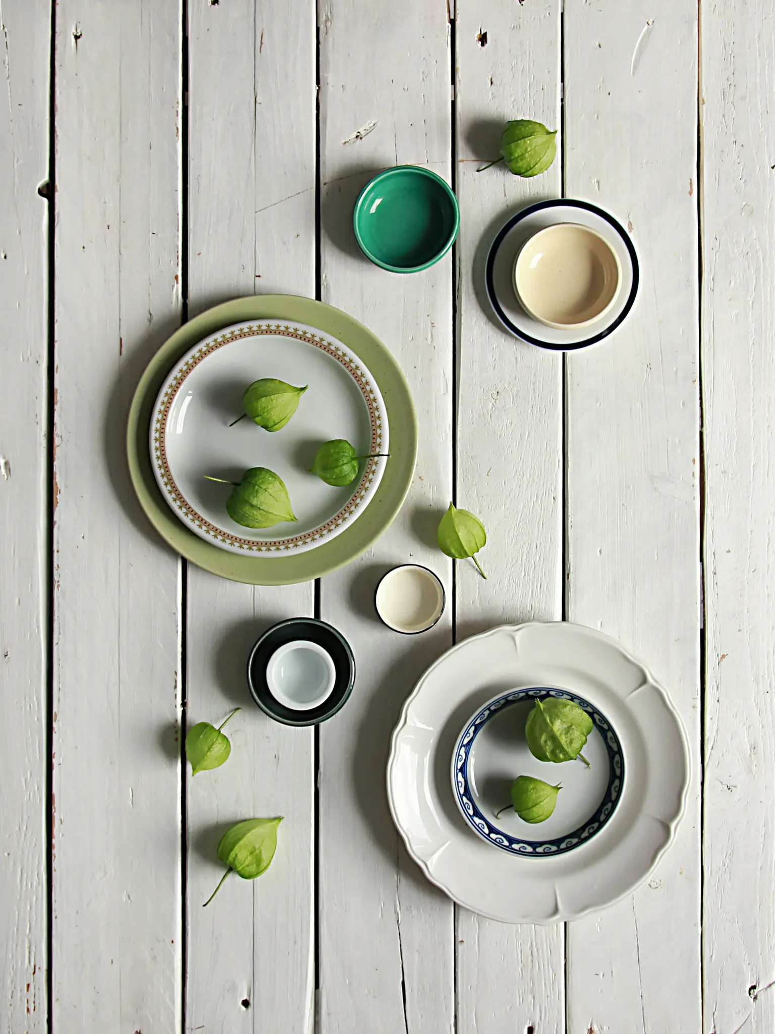 Et overhead fladt lag af grønne tomatillos spredt blandt forskellige keramiske tallerkener og skåle på en slidt hvid