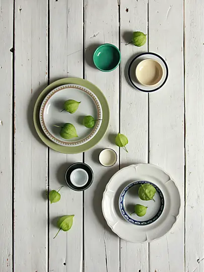 Et overhead fladt lag af grønne tomatillos spredt blandt forskellige keramiske tallerkener og skåle på en slidt hvid