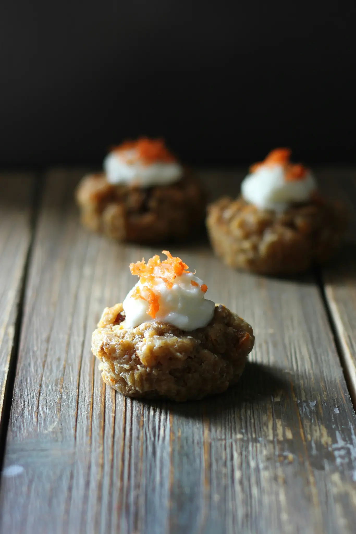 Three small, round baked goods, possibly muffins or cookies, topped with white frosting and shredded orange garnish, on a…