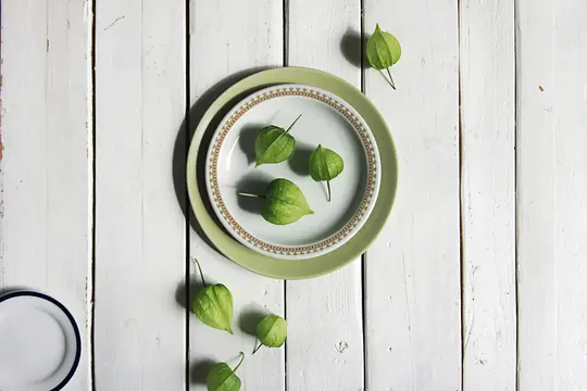 An overhead shot shows several green tomatillos, some on two stacked plates and others scattered on a white wooden surface.
