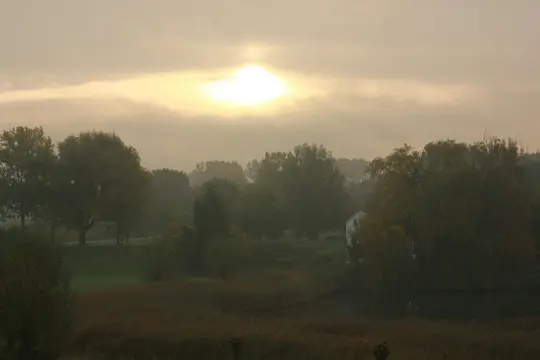 Et bredt landskabsfoto, der viser en diset scene med en klar sol bag skyer, der kaster en glød over træer, et vandområde…