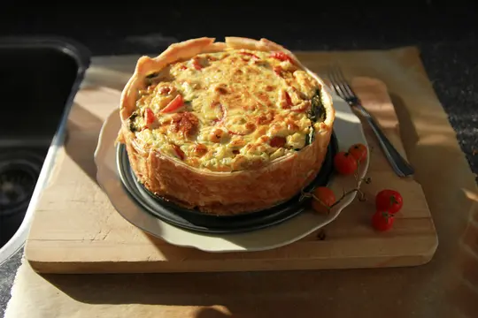 A golden-crusted quiche with a cheesy, egg, tomato, and spinach filling sits on a white plate on a wooden board.