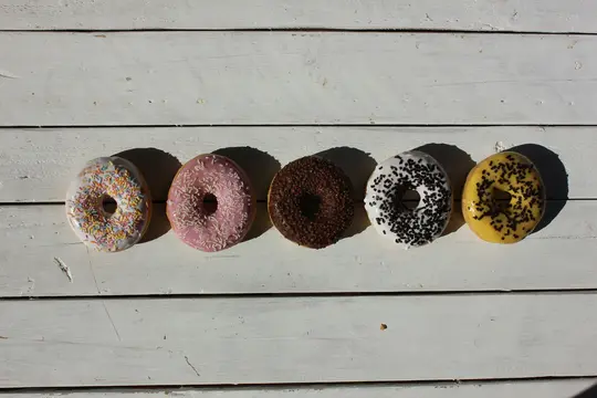 Fem donuts i en række på en hvid træoverflade. Fra venstre mod højre: hvid glasur med farverigt krymmel, lyserød glasur med…