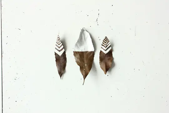 Three dried brown leaves are arranged vertically on a distressed white surface. The middle leaf is half painted white. The…