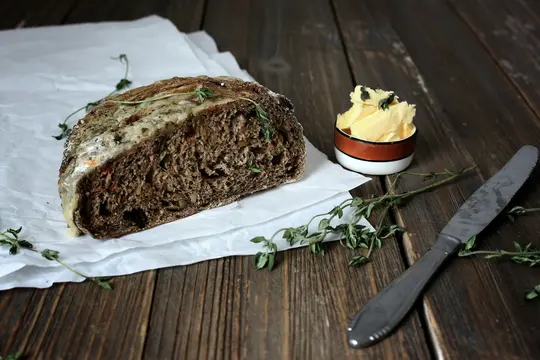 A half-loaf of dark, seeded bread with a light crust sits on white parchment paper on a dark wooden table. A small bowl of…