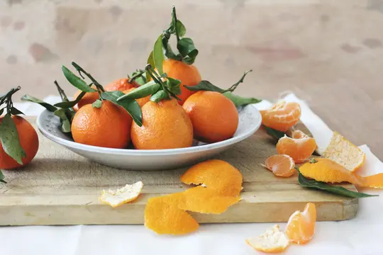 A white bowl holds several whole tangerines with green leaves and stems. Around it, peeled tangerine segments and peels are…