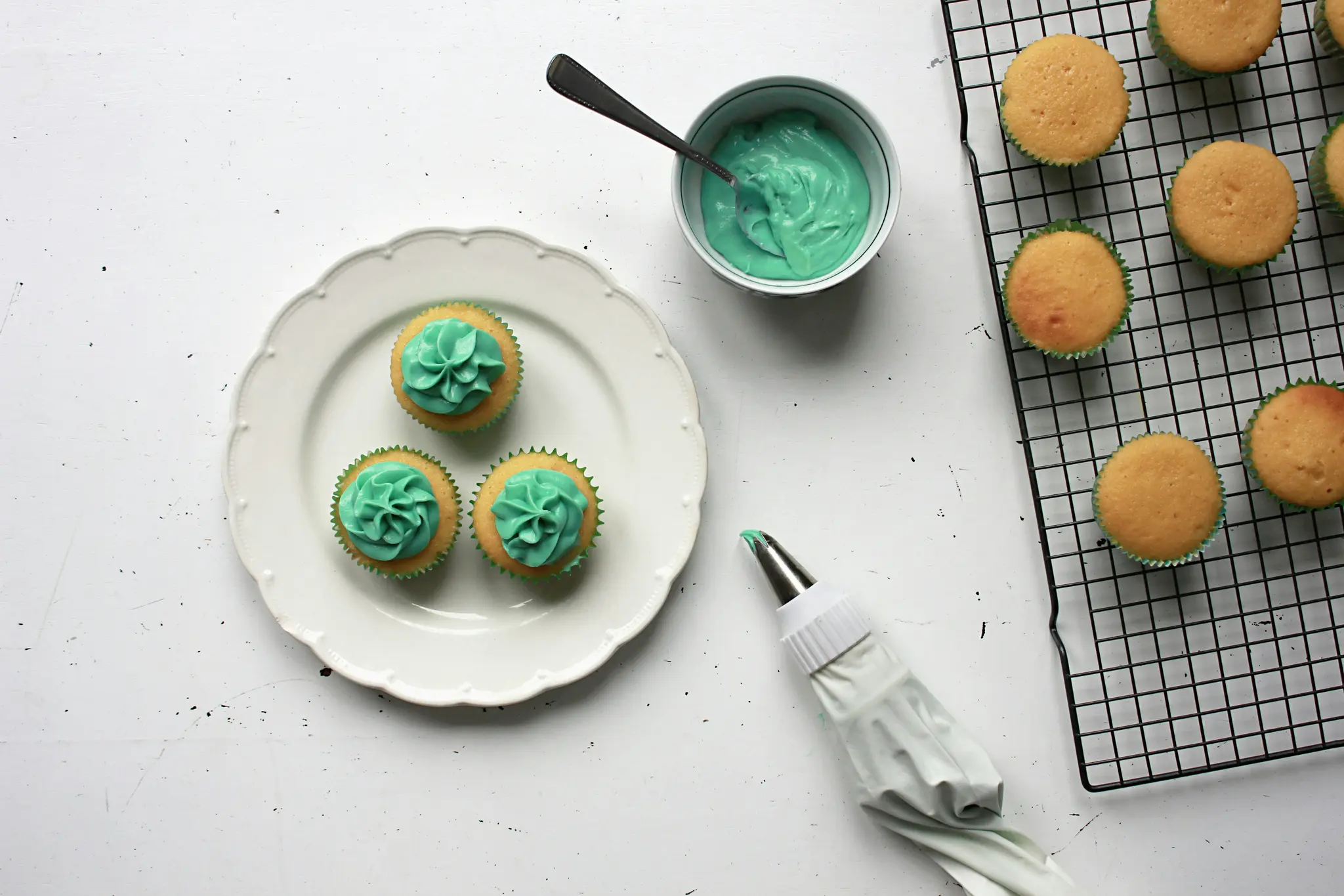 An overhead flat lay shows three cupcakes with teal frosting on a white plate, a bowl of teal frosting, a piping bag, and…