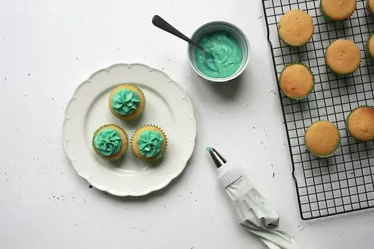 An overhead flat lay shows three cupcakes with teal frosting on a white plate, a bowl of teal frosting, a piping bag, and…
