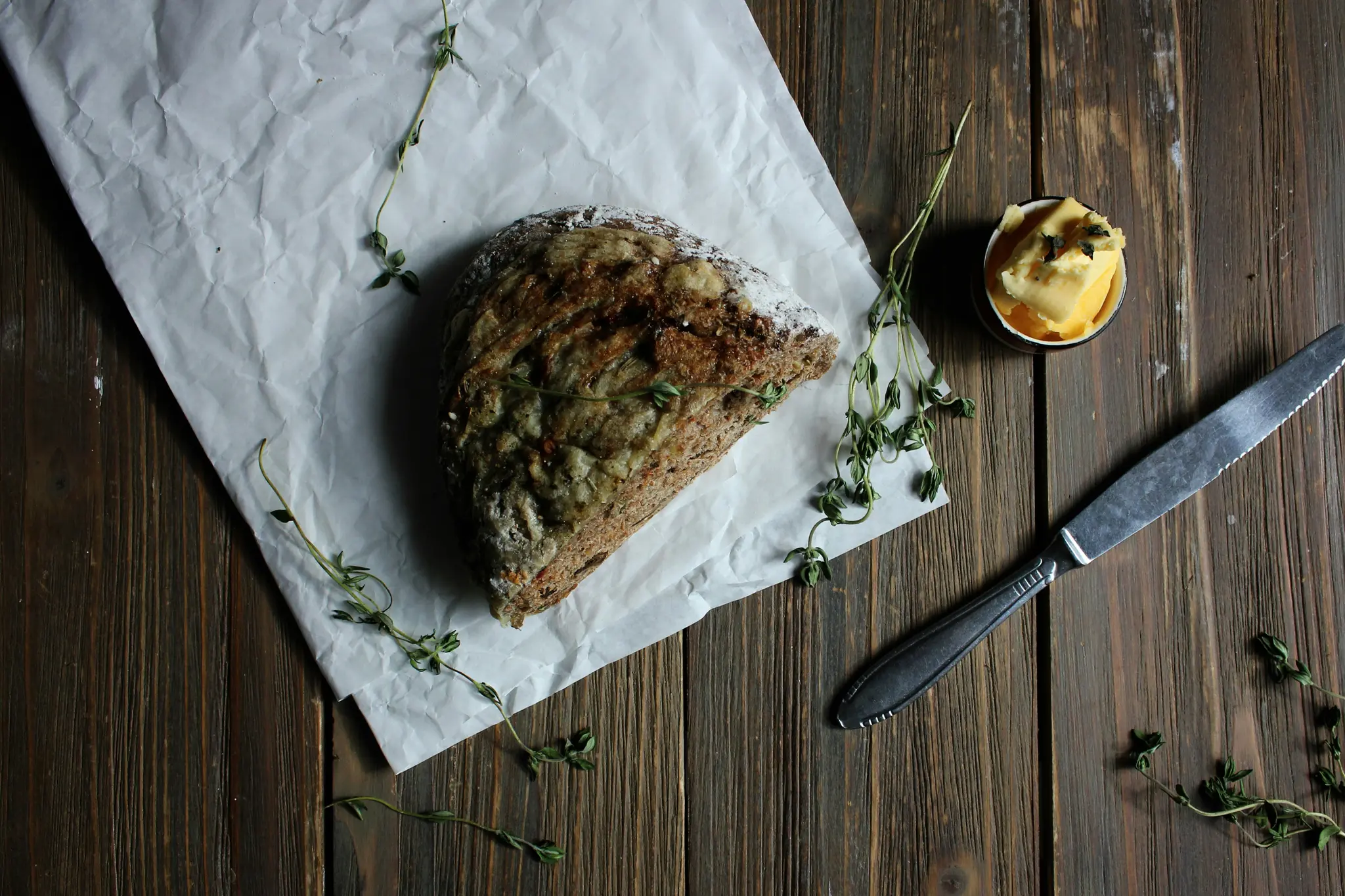 An overhead flat lay shows a half loaf of rustic bread on crumpled white paper, a small bowl of butter, and a knife on a…