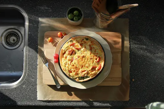 An overhead shot of a golden-brown quiche with visible tomatoes and spinach, on a white plate on a wooden board. A fork…