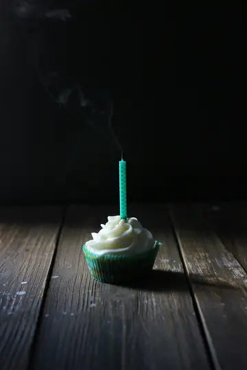Et nærbillede af en cupcake med hvid glasur og et teal-farvet lys, der lige er blevet udblæst, med røg der stiger op.