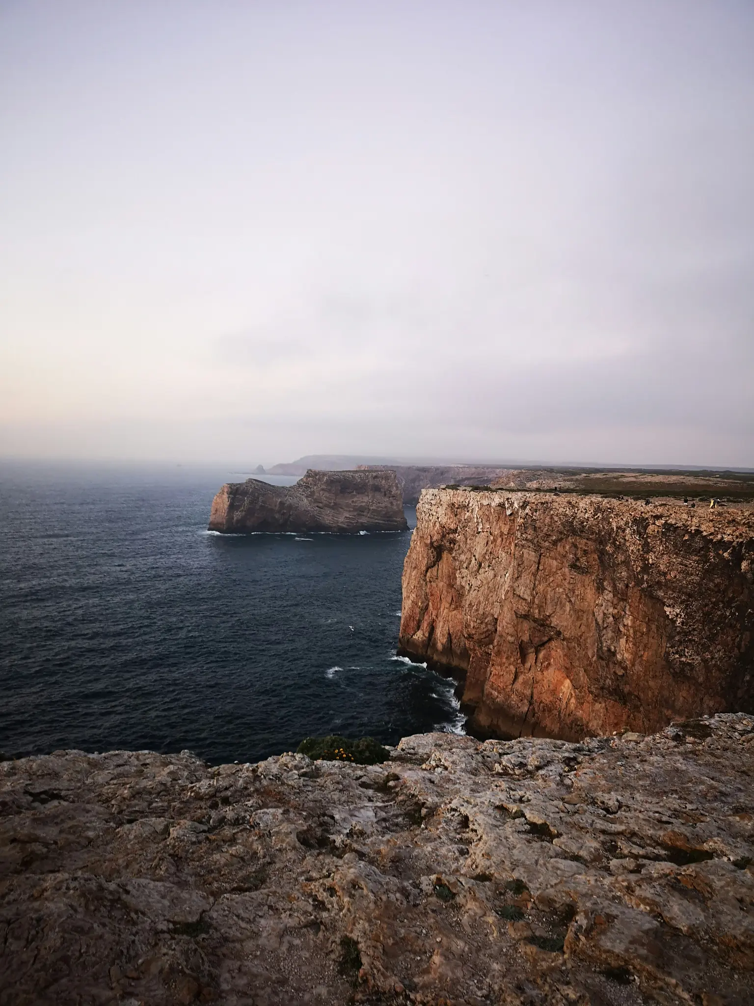 A wide landscape view of rugged, reddish-brown sea cliffs plunging into a dark blue ocean under a vast, grey sky.