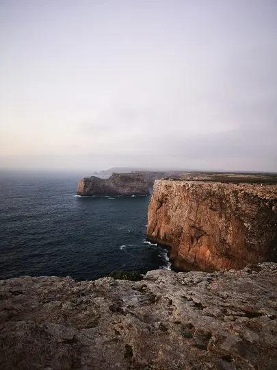 A wide landscape view of rugged, reddish-brown sea cliffs plunging into a dark blue ocean under a vast, grey sky.