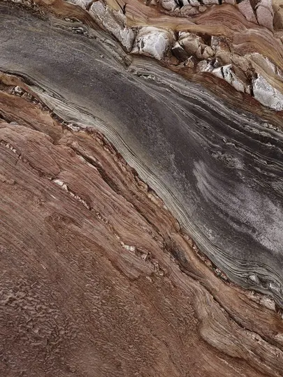 A close-up, abstract view of layered rock formations. The rock shows reddish-brown, dark gray, and white striations.