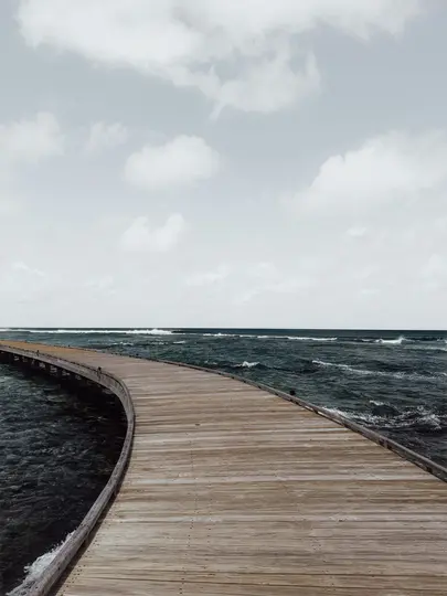 A curved wooden pier stretches into the distance over dark blue ocean water with small waves under a light grey cloudy sky.