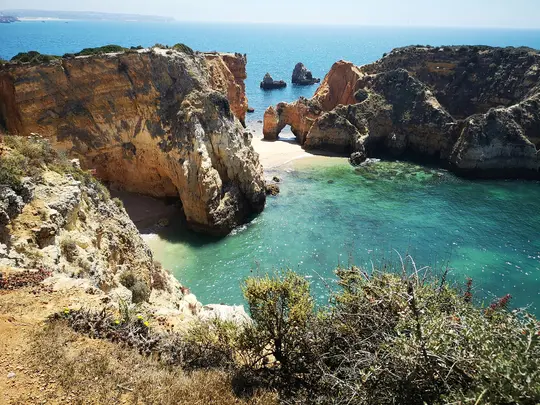 An aerial view of a rugged coastline featuring orange-brown cliffs, small sandy beaches, and clear turquoise ocean water.