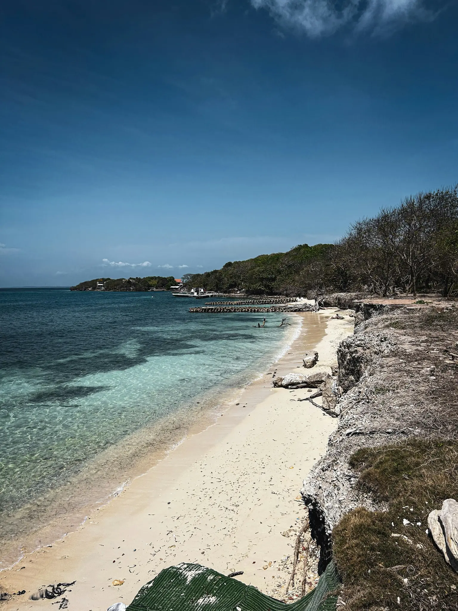 A high-angle view of a tropical beach with clear turquoise water, white sand, and a rocky cliff on the right.