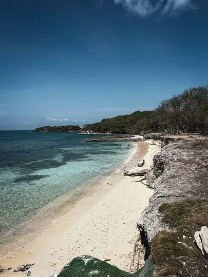 A high-angle view of a tropical beach with clear turquoise water, white sand, and a rocky cliff on the right.