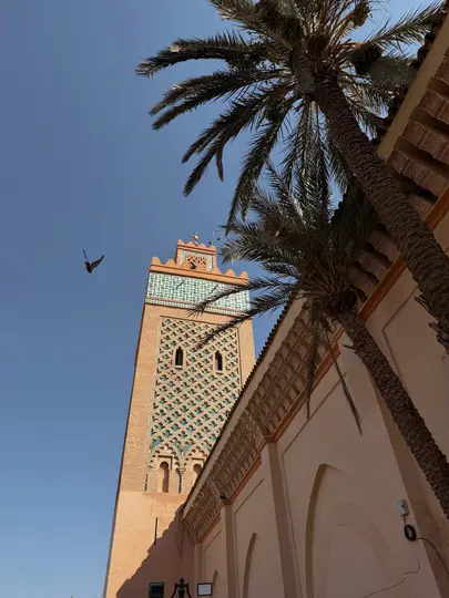 Vista de ángulo bajo de un minarete alto de ladrillo con intrincados patrones turquesa y blancos, enmarcado por dos palmeras.
