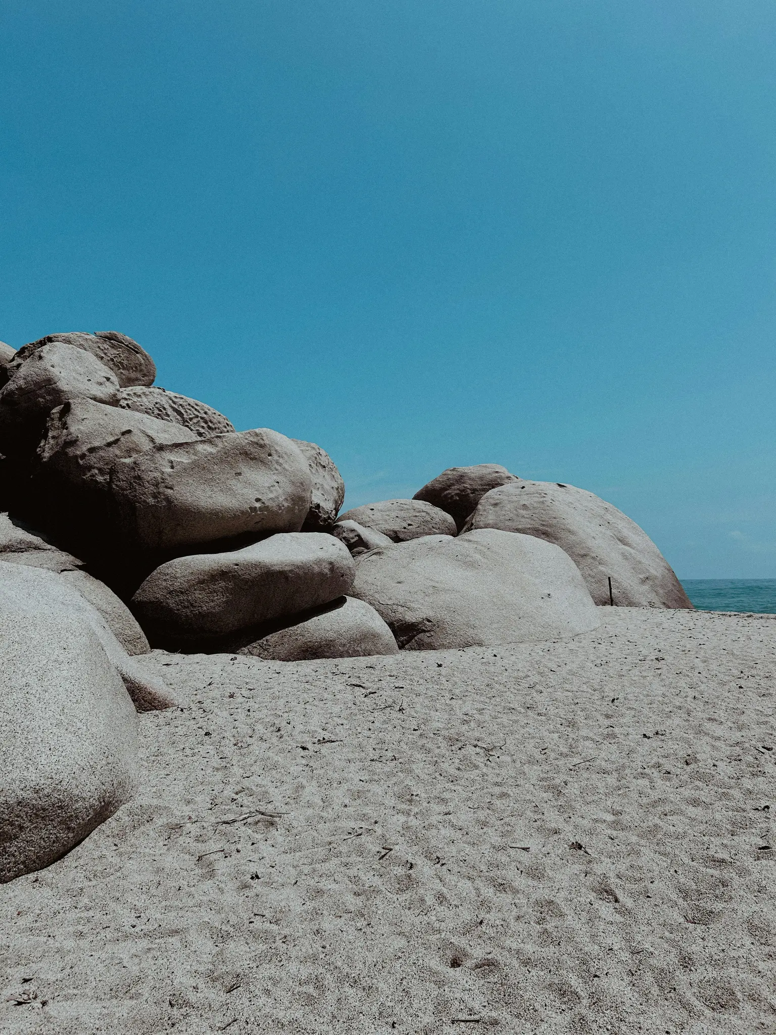 A low-angle shot of a sandy beach with large, smooth, light-colored boulders on the left and center. A clear blue sky…
