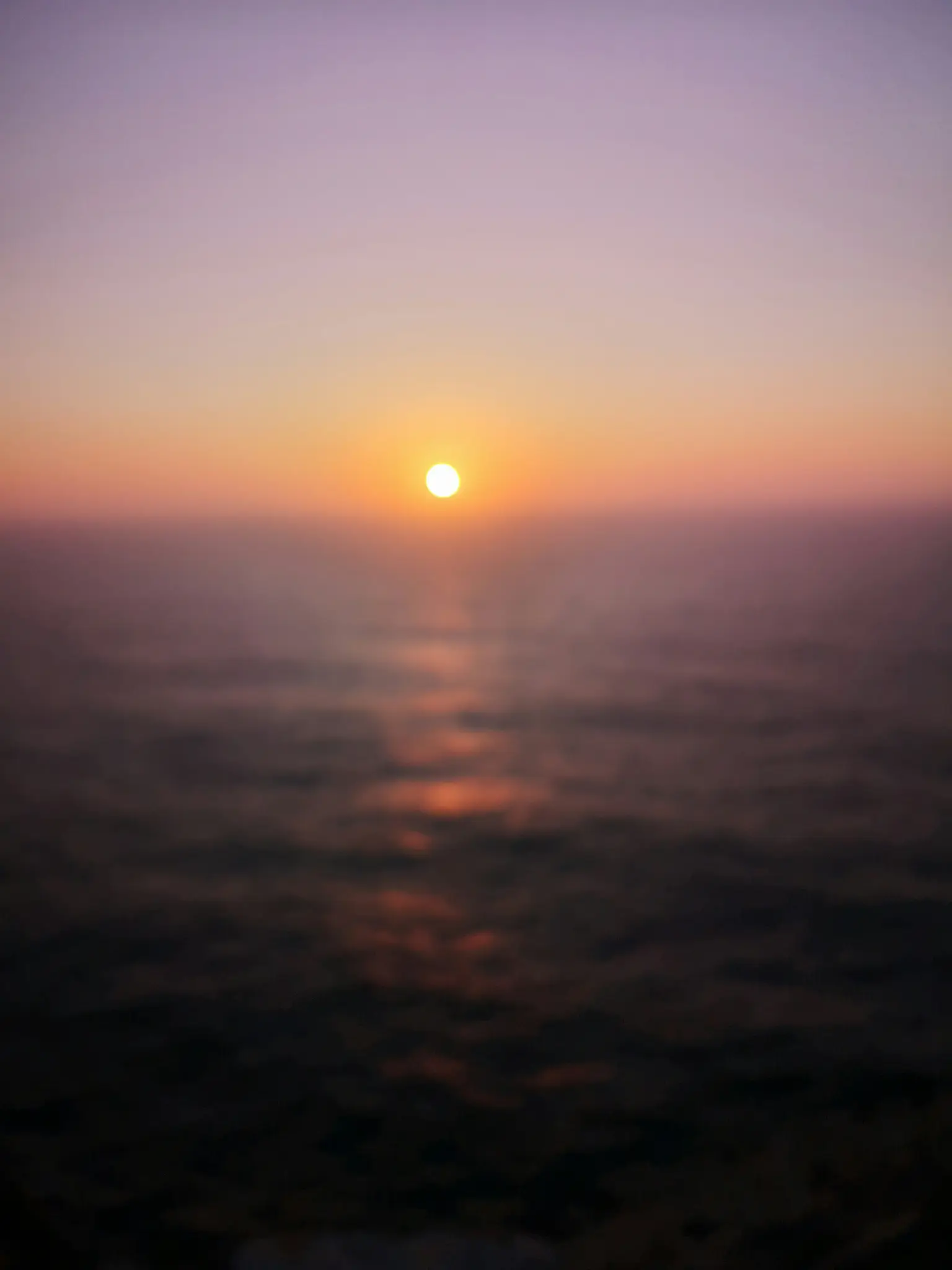 A blurred photo of a sunset over a body of water. The sun is a bright orange circle near the horizon, casting a shimmering…