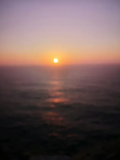 A blurred photo of a sunset over a body of water. The sun is a bright orange circle near the horizon, casting a shimmering…