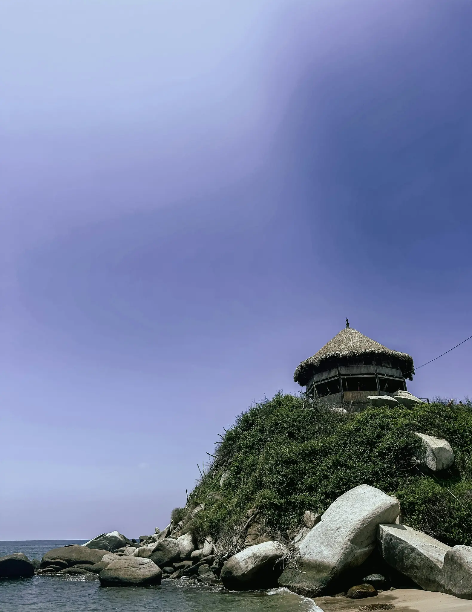 A round, two-story building with a conical thatched roof sits on a verdant hill. Large boulders line the sandy beach and…