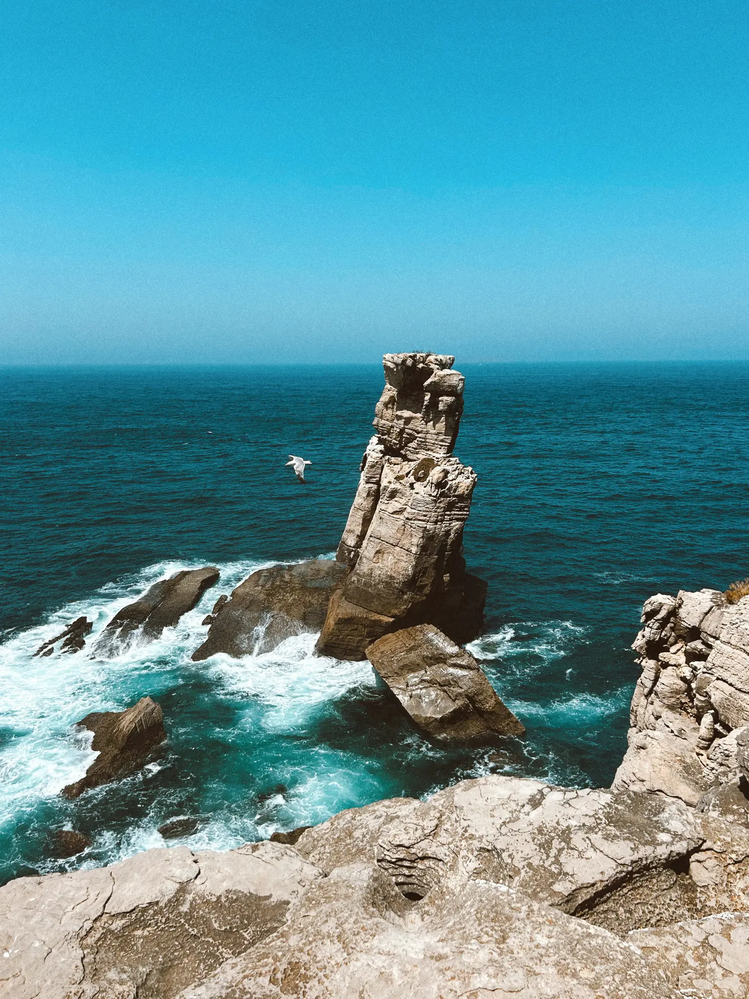 Un farallón alto y escarpado se eleva desde el océano turquesa, rodeado de olas blancas y rompientes. Un pájaro vuela en el