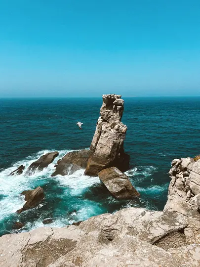 Un farallón alto y escarpado se eleva desde el océano turquesa, rodeado de olas blancas y rompientes. Un pájaro vuela en el