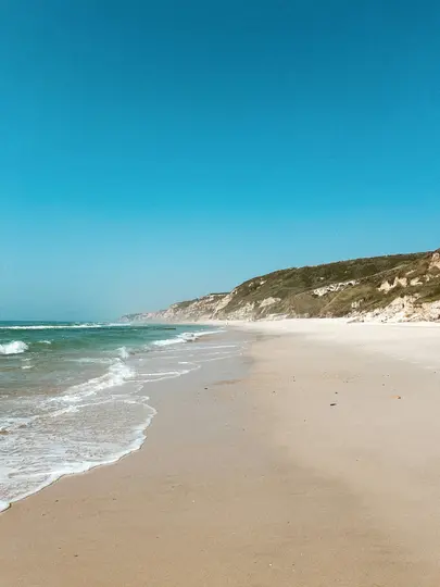 A wide-angle view of a sandy beach with gentle waves breaking on the shore and cliffs covered in green vegetation.