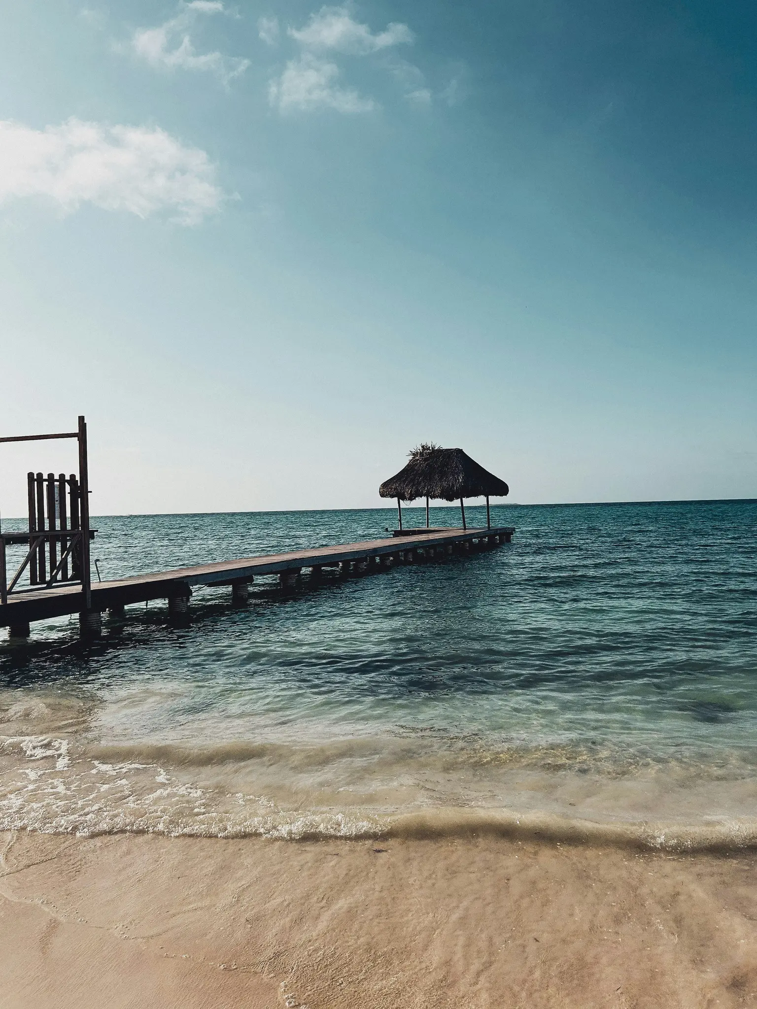 A long wooden pier with a small thatched-roof hut at its end stretches into calm turquoise ocean water from a sandy beach.