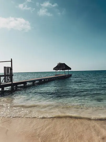 A long wooden pier with a small thatched-roof hut at its end stretches into calm turquoise ocean water from a sandy beach.