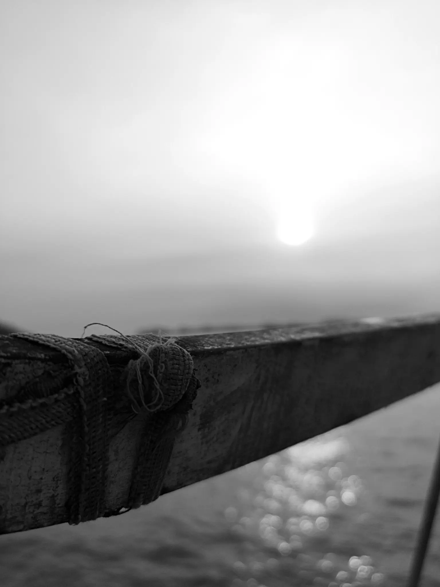 A black and white close-up of a wooden boat's edge, wrapped with rope, against a bright sun over shimmering water.