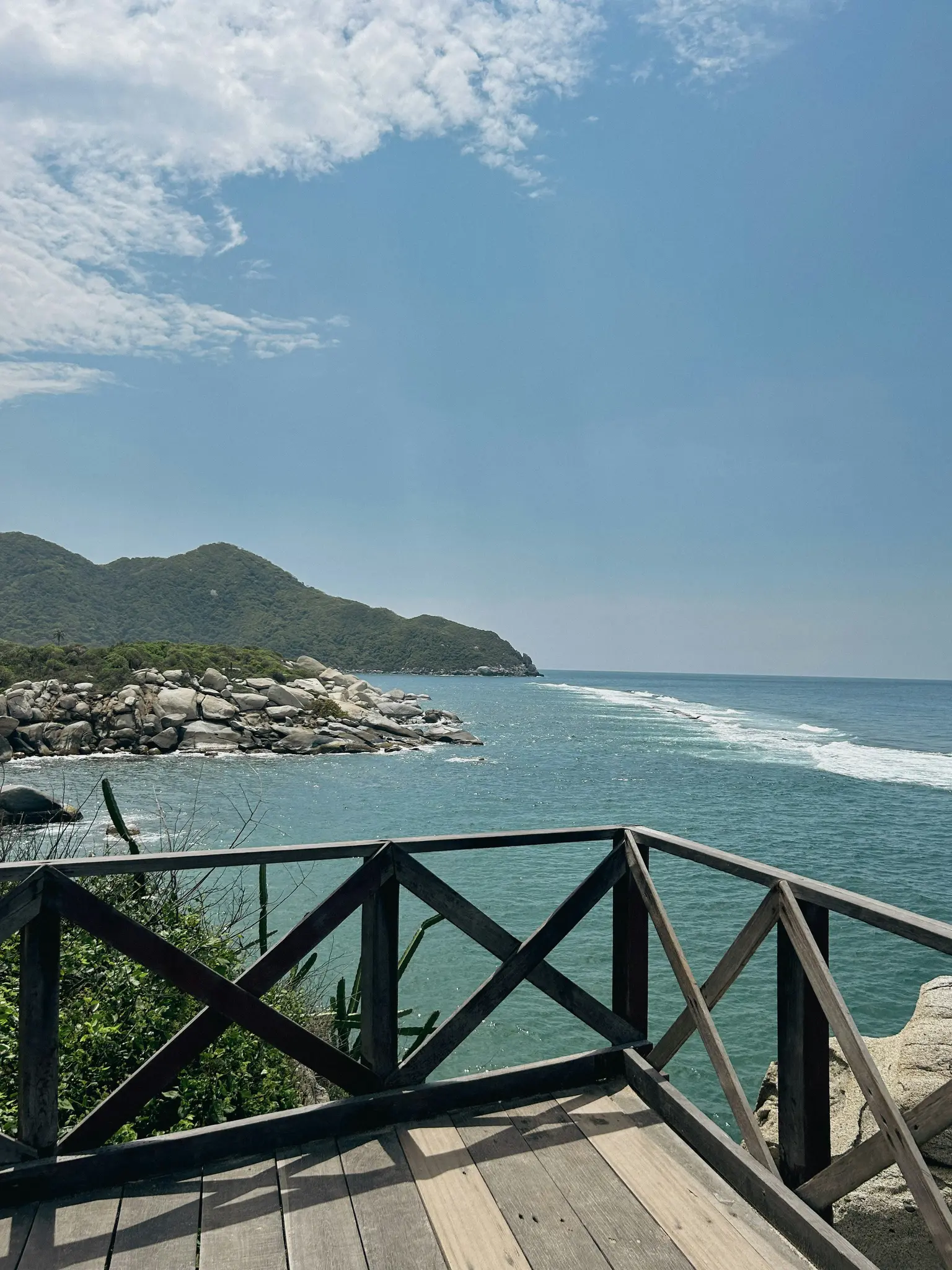 A wooden deck with a criss-cross railing overlooks a turquoise ocean with white waves, a rocky coastline, and green hills.