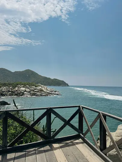 A wooden deck with a criss-cross railing overlooks a turquoise ocean with white waves, a rocky coastline, and green hills.