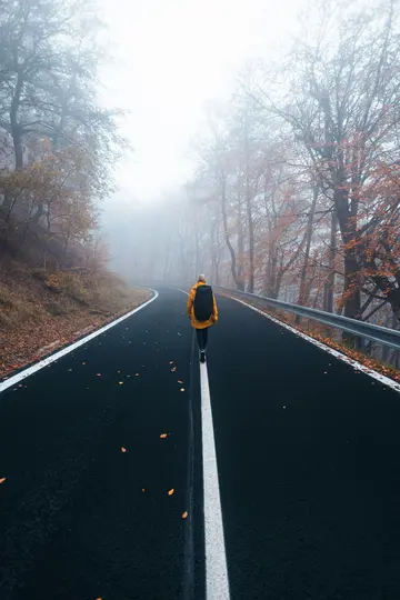 Una persona con una chaqueta amarilla brillante y una mochila negra camina por la línea blanca de una carretera asfaltada