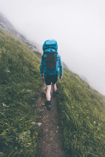 Eine Person mit einem großen blauen Rucksack geht auf einem schmalen Pfad einen steilen, grasbewachsenen Berghang hinunter im