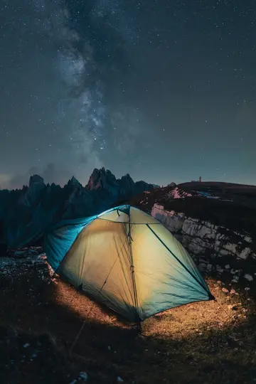 Ein leuchtend gelbes und blaues Zelt leuchtet von innen auf einem dunklen, felsigen Berghang unter einem klaren Nachthimmel
