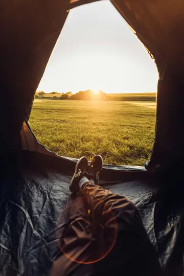 Eine Ansicht von innen eines dunklen Zeltes, die auf ein lebendiges grünes Feld blickt, das im warmen Licht einer aufgehenden