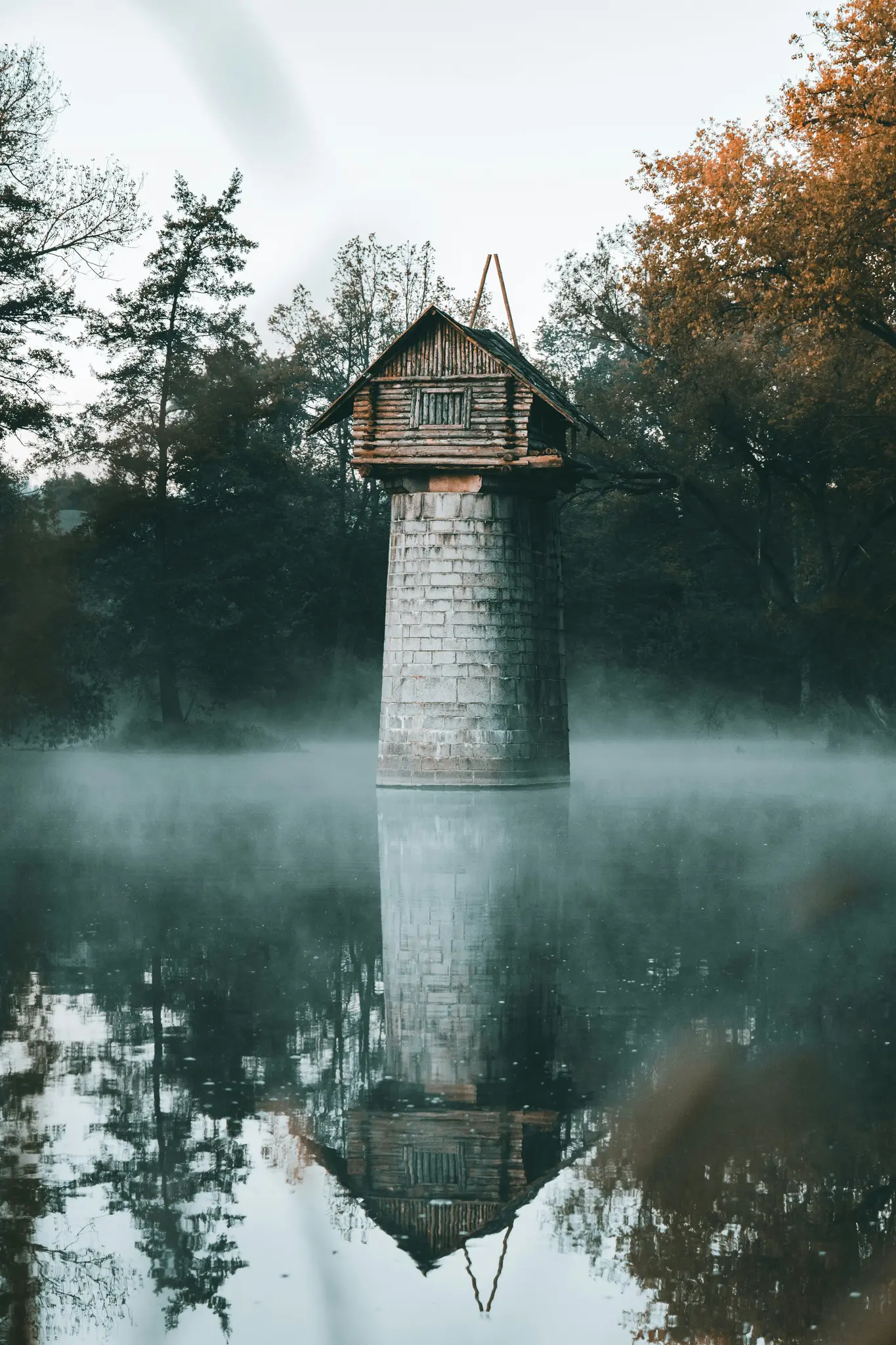 Ein kleines, rustikales Holzhaus mit Satteldach steht auf einem hohen, zylindrischen Steinturm in einem nebligen See. Bäume