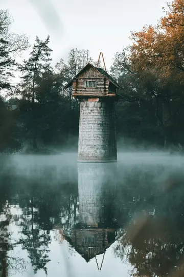 Una pequeña y rústica casa de madera con techo a dos aguas se asienta sobre una torre alta y cilíndrica de piedra en un lago