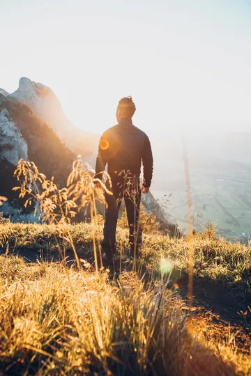 Eine Person steht auf einem Berggrat, mit dem Rücken zum Betrachter, und blickt auf ein sonnenbeschienenes Tal und Berge.