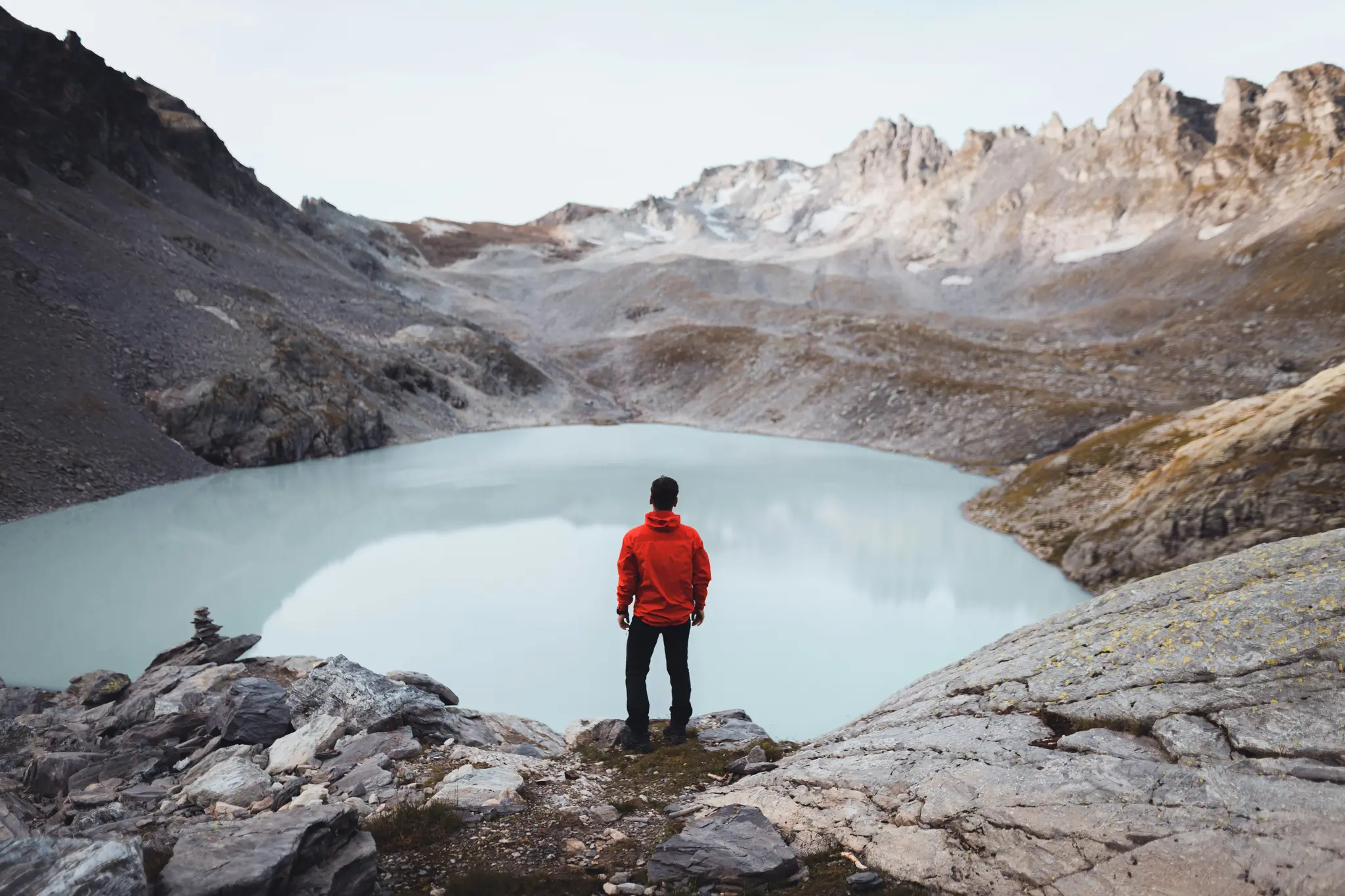 Una persona con una chaqueta roja brillante se encuentra en una repisa rocosa, contemplando un lago glaciar tranquilo y de