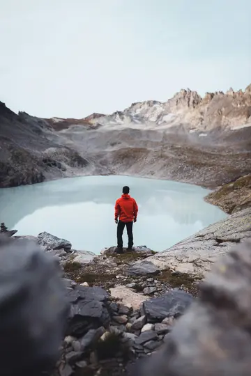 Una persona con una chaqueta roja brillante se para en una repisa rocosa, mirando un lago glaciar tranquilo de color azul