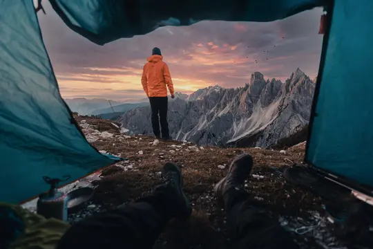 Eine Person in einer orangefarbenen Jacke steht auf einem Bergrücken und blickt auf eine zerklüftete Bergkette unter einem