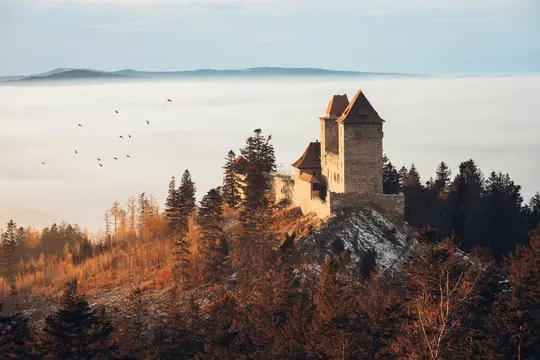 Eine Steinfestung mit roten Dächern thront auf einem Hügel mit Herbstbäumen und Schneefeldern und blickt auf ein Nebelmeer.