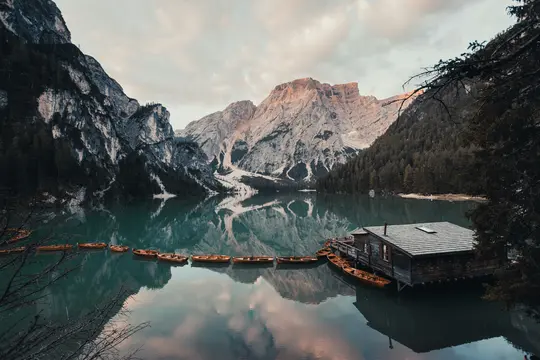 Una amplia foto paisajística de un lago de montaña tranquilo con un embarcadero de madera y varios botes de remo. Montañas