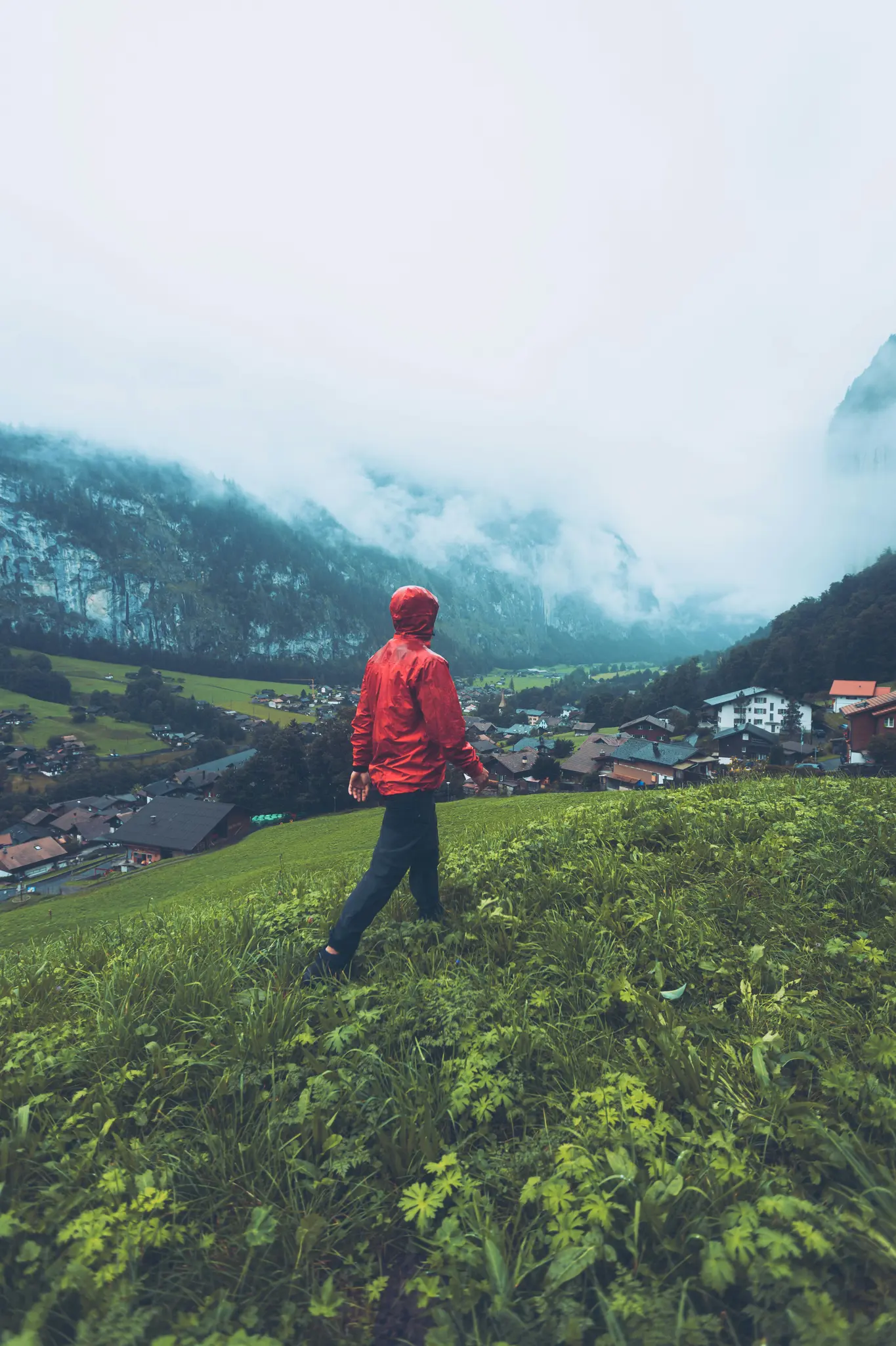 Eine Person in einer leuchtend roten Regenjacke geht einen üppigen grünen Hang hinunter und blickt auf ein Dorf in einem