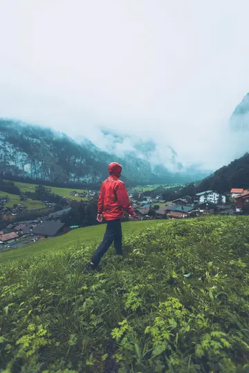 Eine Person in einer leuchtend roten Regenjacke geht einen üppigen grünen Hang hinunter und blickt auf ein Dorf in einem