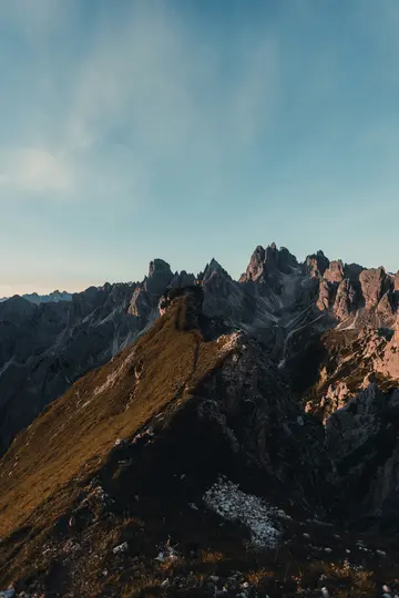 Eine weite Landschaftsaufnahme einer Bergkette während der goldenen Stunde. Ein grasbewachsener Kamm führt zu zerklüfteten